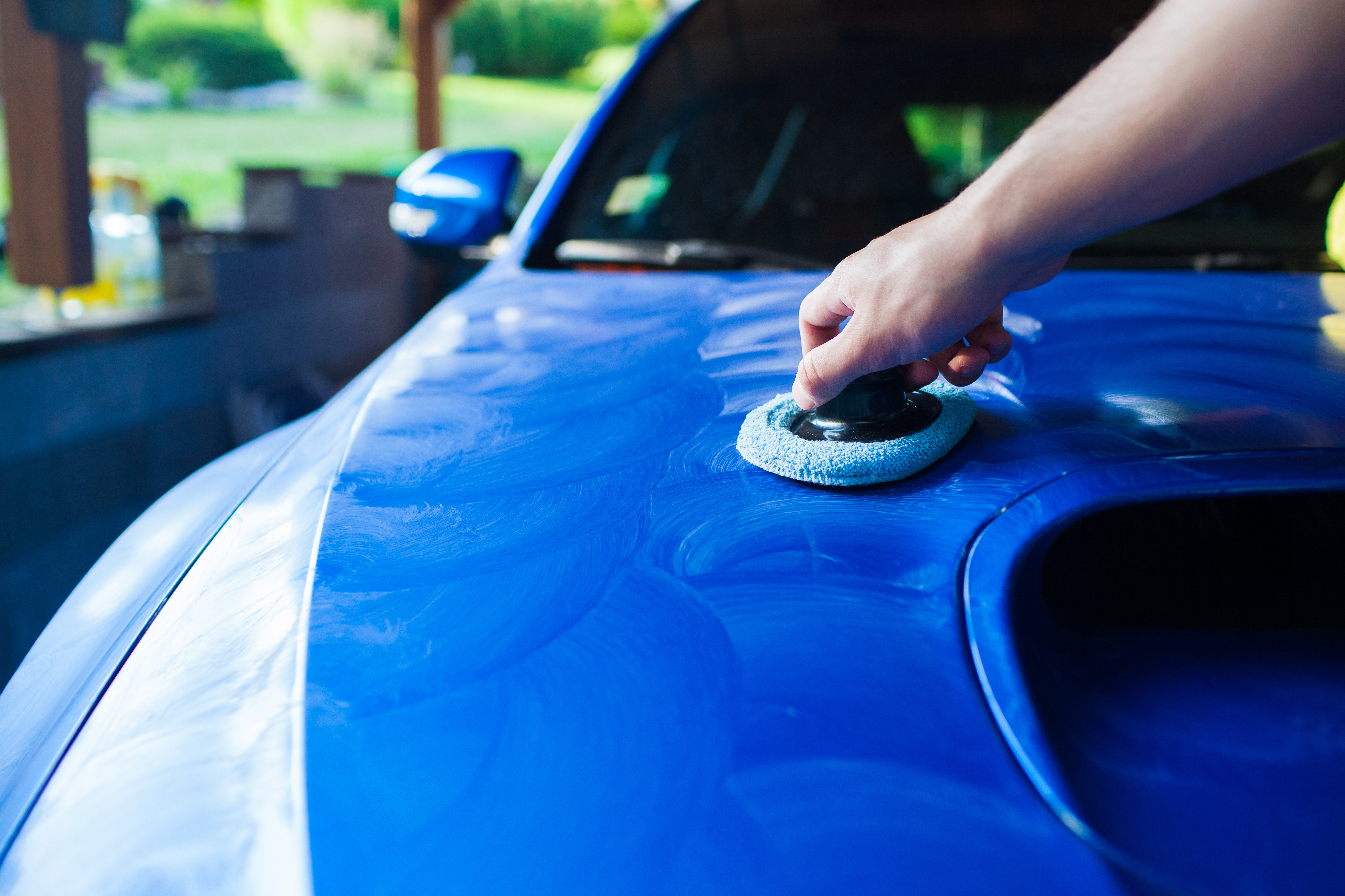 Person using gray microfiber pad to polish blue car hood outdoors